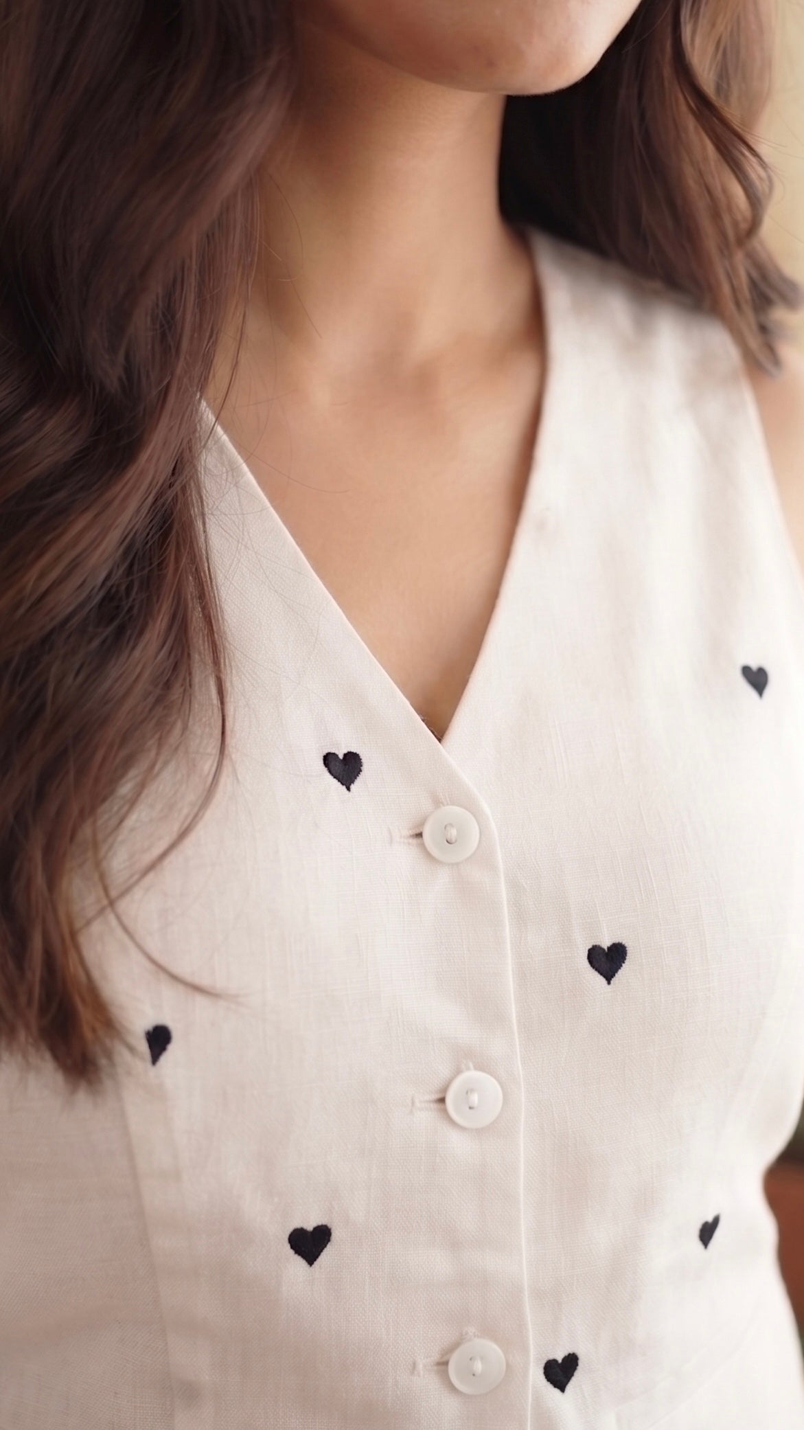 A close-up of a woman wearing a sleeveless white V-neck top with small black heart embroidery and white buttons, photographed in a softly lit indoor setting.
