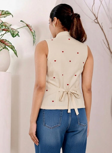 Woman wearing a beige sleeveless top with heart patterns and blue jeans, standing against a light background.