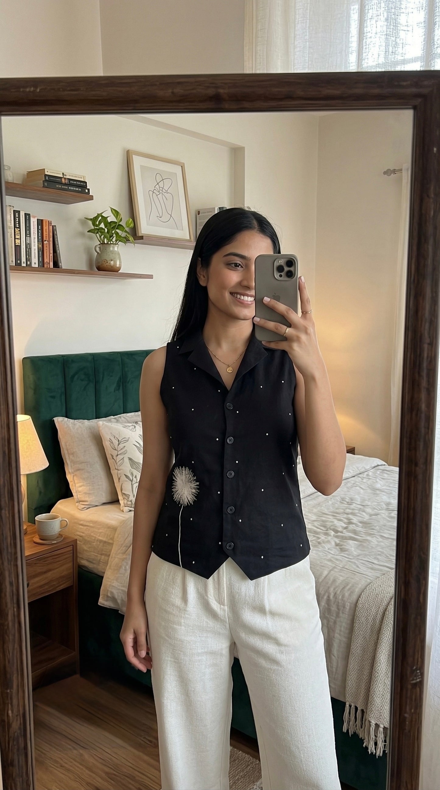 Woman taking a mirror selfie in a bedroom wearing a black sleeveless top and white pants.