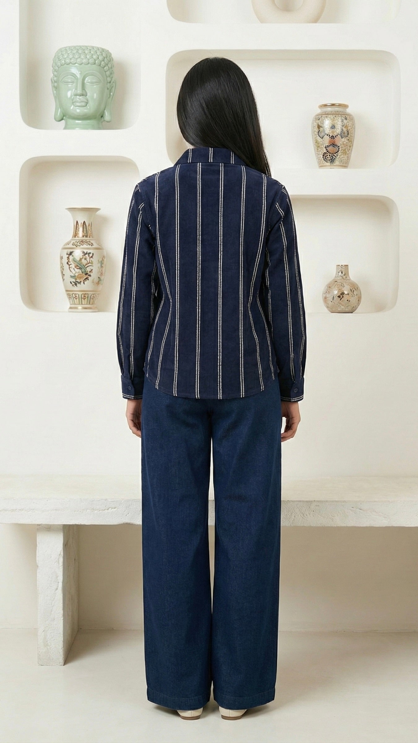Person wearing a navy pinstripe blazer and wide-leg pants in a room with decorative vases on shelves.