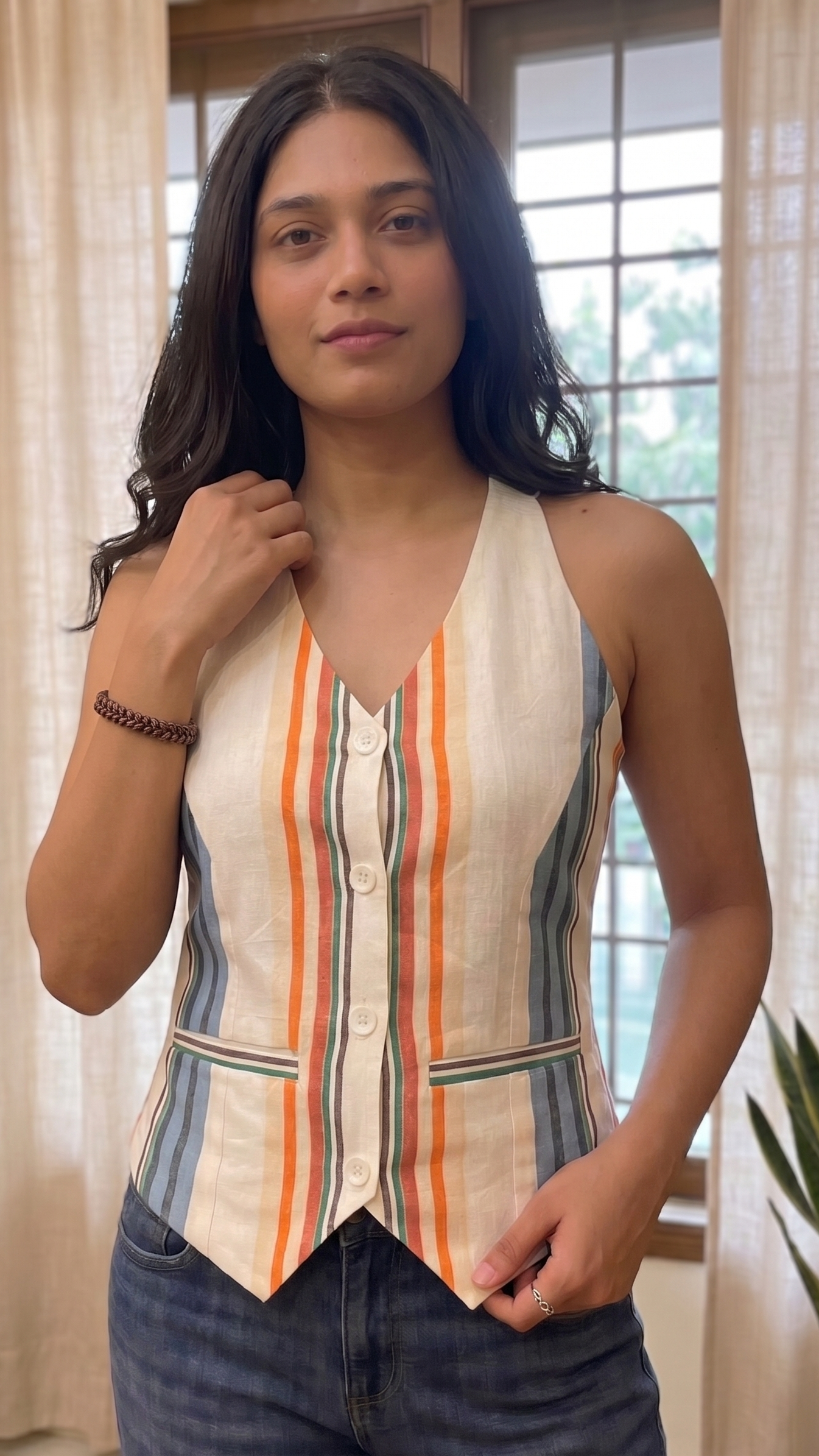 Woman wearing a sleeveless striped top indoors with curtains and plants in the background
