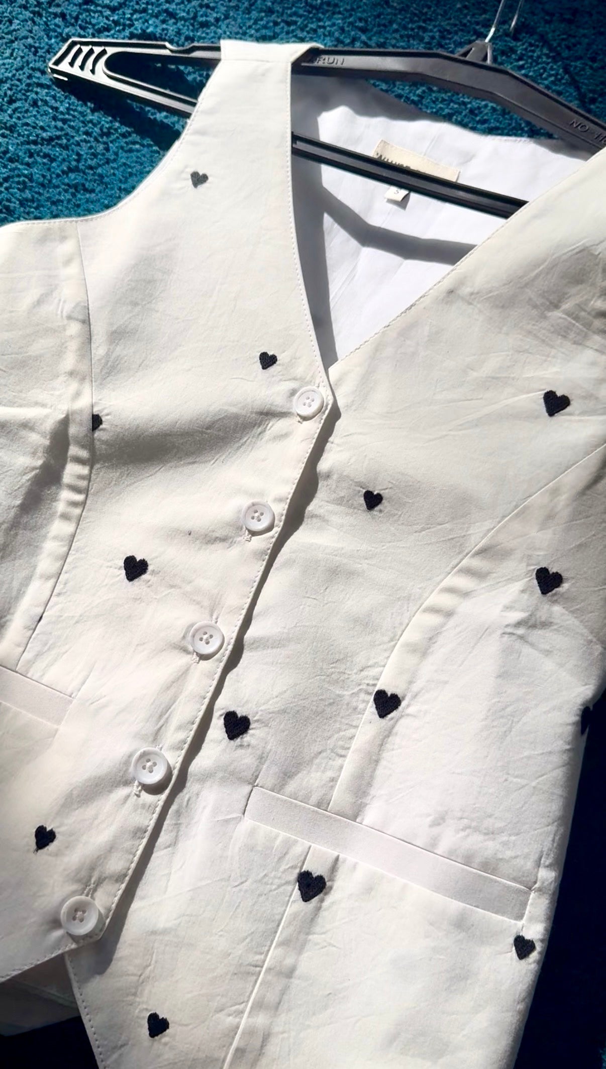 A white sleeveless vest with small black heart embroidery, shown hanging on a black hanger against a textured blue background.