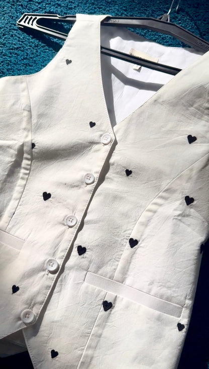 A white sleeveless vest with small black heart embroidery, shown hanging on a black hanger against a textured blue background.