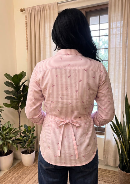 Person wearing a pink patterned shirt with a belt in a room with plants and a window.