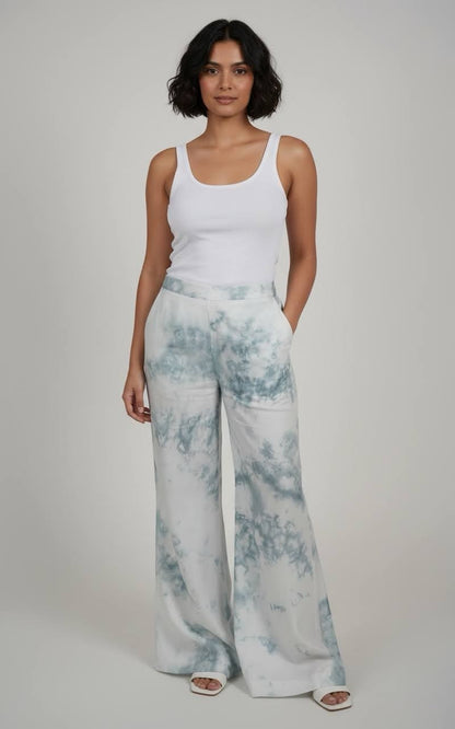 Woman wearing a white tank top and light blue tie-dye pants on a plain background