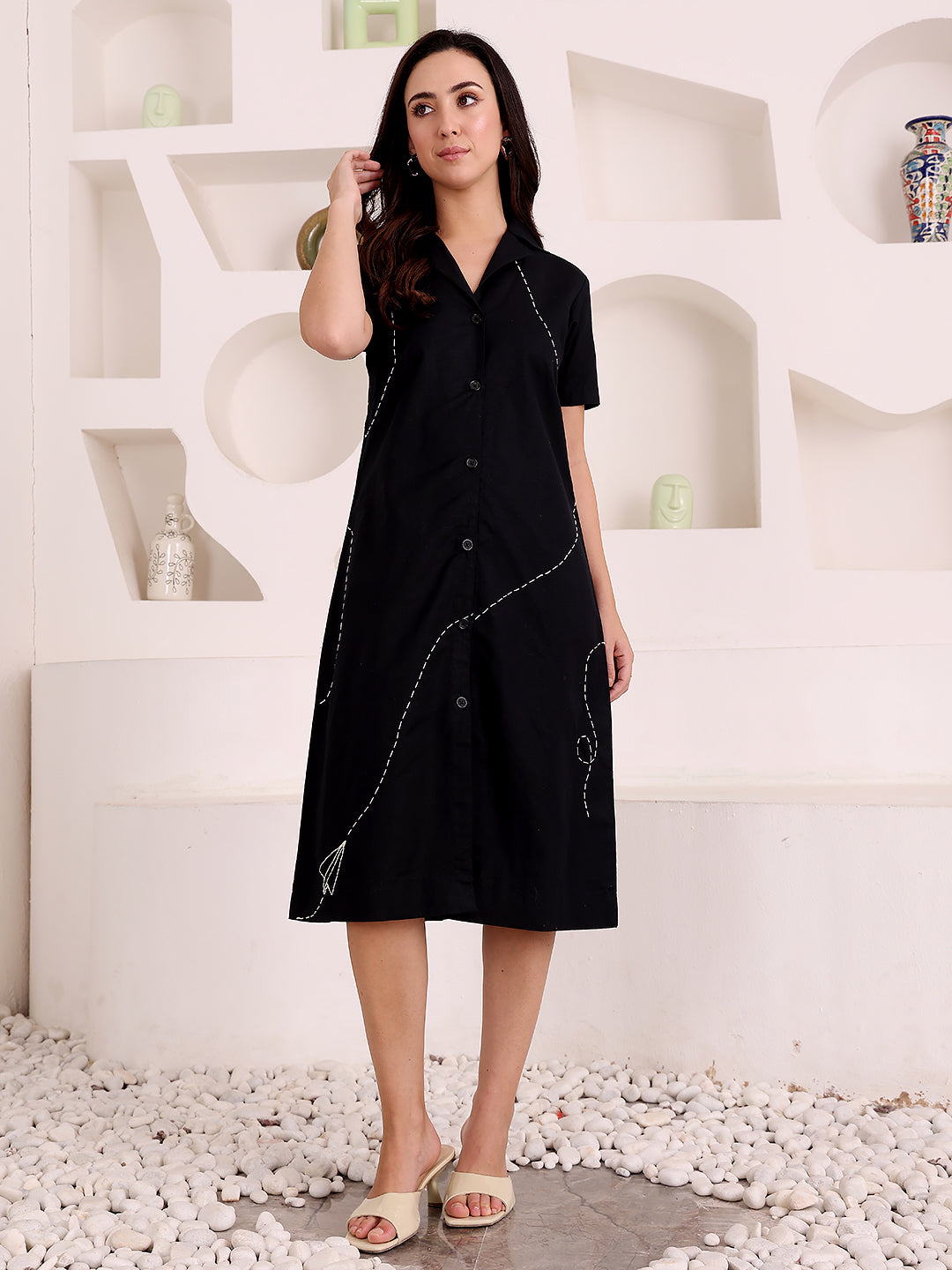 Woman wearing a black dress with white embroidery in a minimalistic setting.
