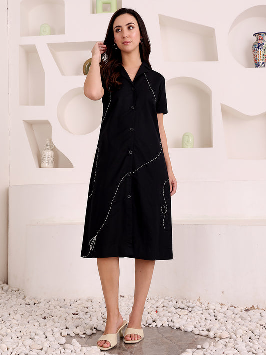 Woman wearing a black dress with white embroidery in a minimalistic setting.
