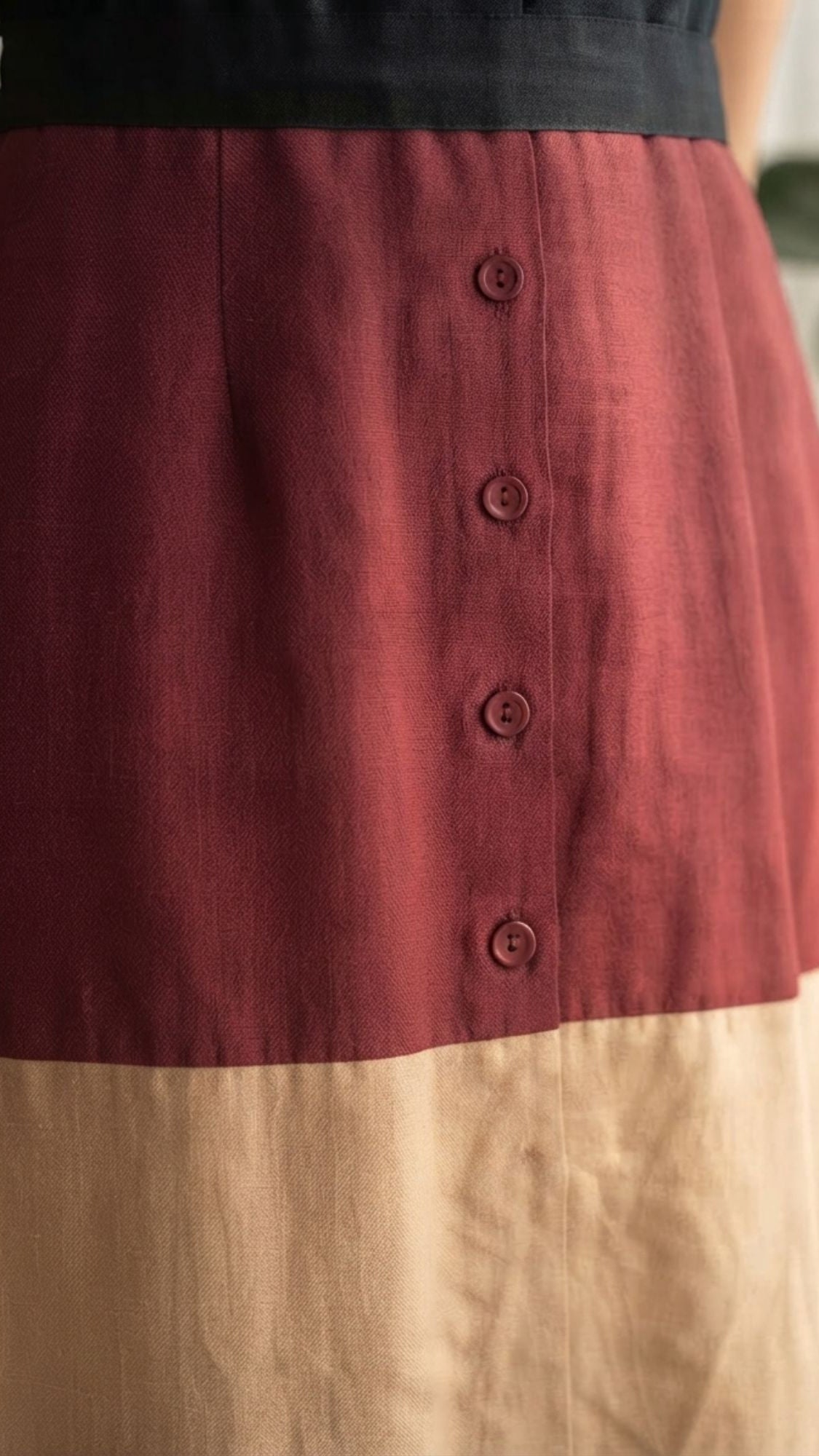 A close-up of a color-blocked skirt with a deep red upper section featuring a vertical button placket and a light beige lower section, shown on a model.