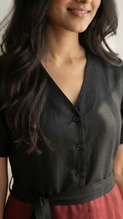 A woman wearing a black short-sleeve linen button-front top with a tie waist, paired with a rust-red skirt, in a softly lit indoor setting.